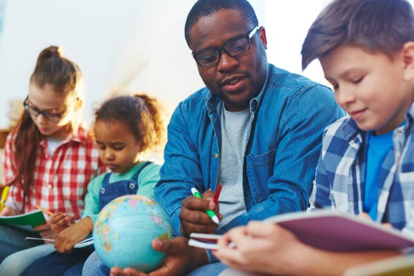 Teacher helping three students learn while he holds a globe and points to a section of a book.